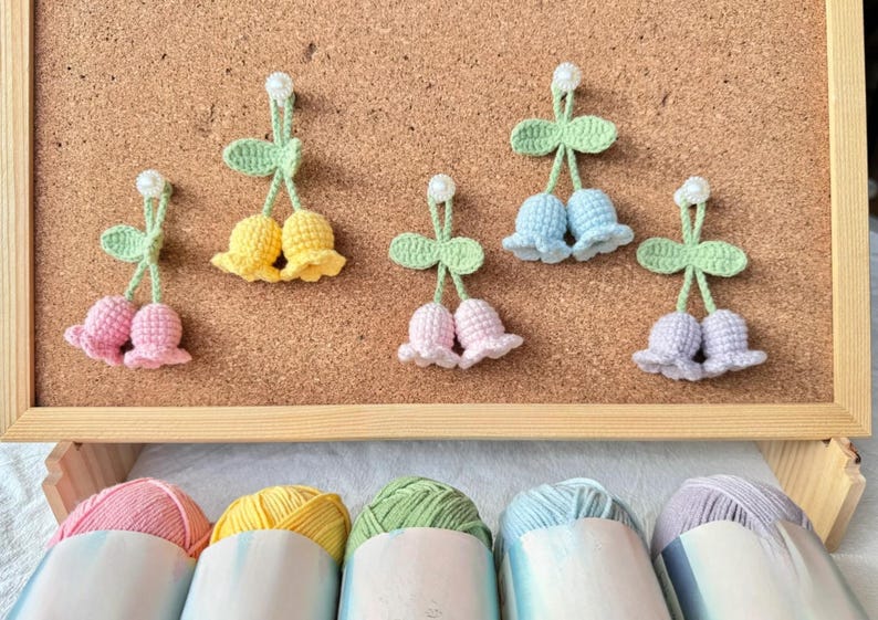May include: Crocheted tulip decorations in pink, yellow, blue, and lavender, attached to a corkboard with push pins. The tulips have green stems and leaves. Below are rolls of yarn in matching colors.