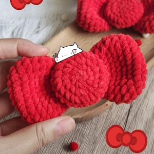 May include: Close-up of three bright red, plush, heart-shaped crochet items. A small white cat illustration is on top of one heart. The hearts are on a wooden tray. A red pom-pom and a red bow illustration are also visible.