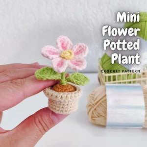 May include: A miniature crochet flower potted plant with a pink and yellow flower, green leaves, and a beige pot. The text "Mini Flower Potted Plant CROCHET PATTERN" is visible in the image. The plant is held in a hand.