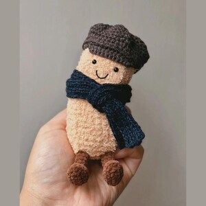 May include: A handmade plush toy shaped like a cylinder, wearing a dark blue scarf and a brown cap. The toy has a smiling face and brown feet. The plushie is held in a person's hand.