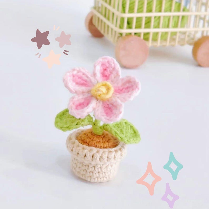 May include: A crocheted flower in a small pot. The flower has pink petals with a yellow centre, green leaves, and sits in a beige pot. The pot is filled with orange yarn, and the background is white.