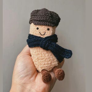 Puede incluir: Un juguete de peluche beige con una cara sonriente, una bufanda azul oscuro y una gorra marrón. El juguete tiene pequeños pies marrones y está diseñado para parecerse a un personaje. El juguete se sostiene en una mano.
