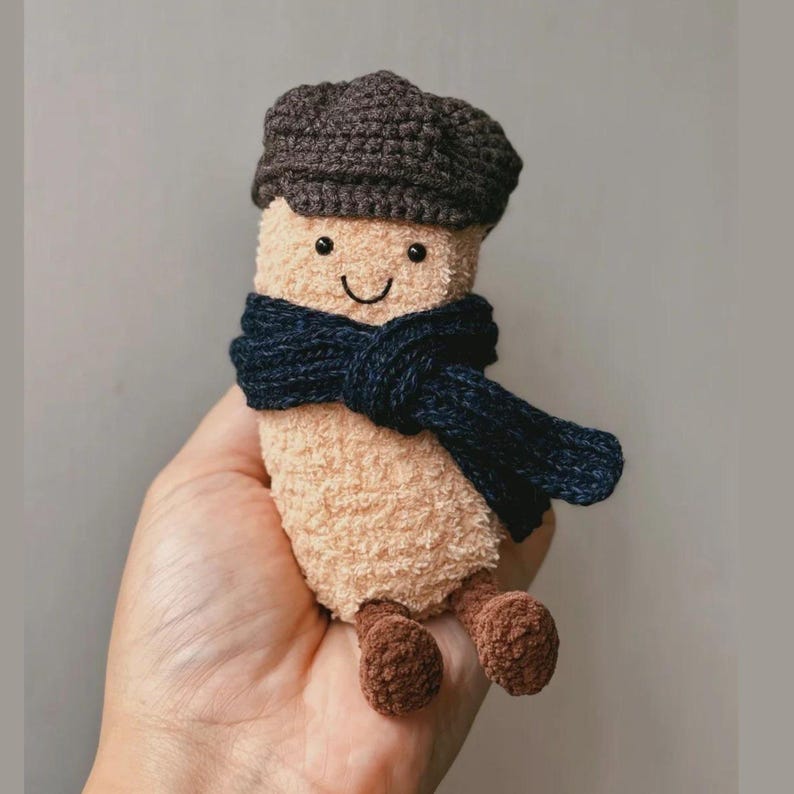May include: A plush, beige-colored toy with a smiling face, wearing a dark blue scarf and a brown cap. The toy has small brown feet and is designed to look like a character. The toy is held in a hand.