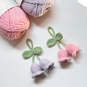 May include: Close-up of two crocheted bellflower ornaments in pink and lavender, each with a green leaf and loop. Two balls of yarn in pink and lavender are in the background. The ornaments are handmade.