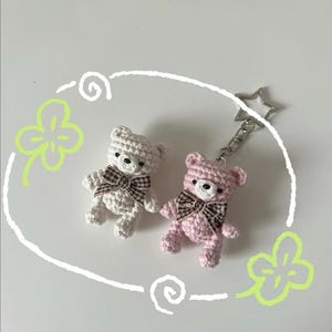 Puede incluir: Dos llaveros de osos de peluche de ganchillo. Uno es blanco, el otro rosa. Ambos tienen lazos a cuadros marrones y ojos negros. Un llavero plateado en forma de estrella est&aacute; unido al oso rosa. Los osos est&aacute;n sobre un fondo blanco con elementos decorativos verdes y blancos.