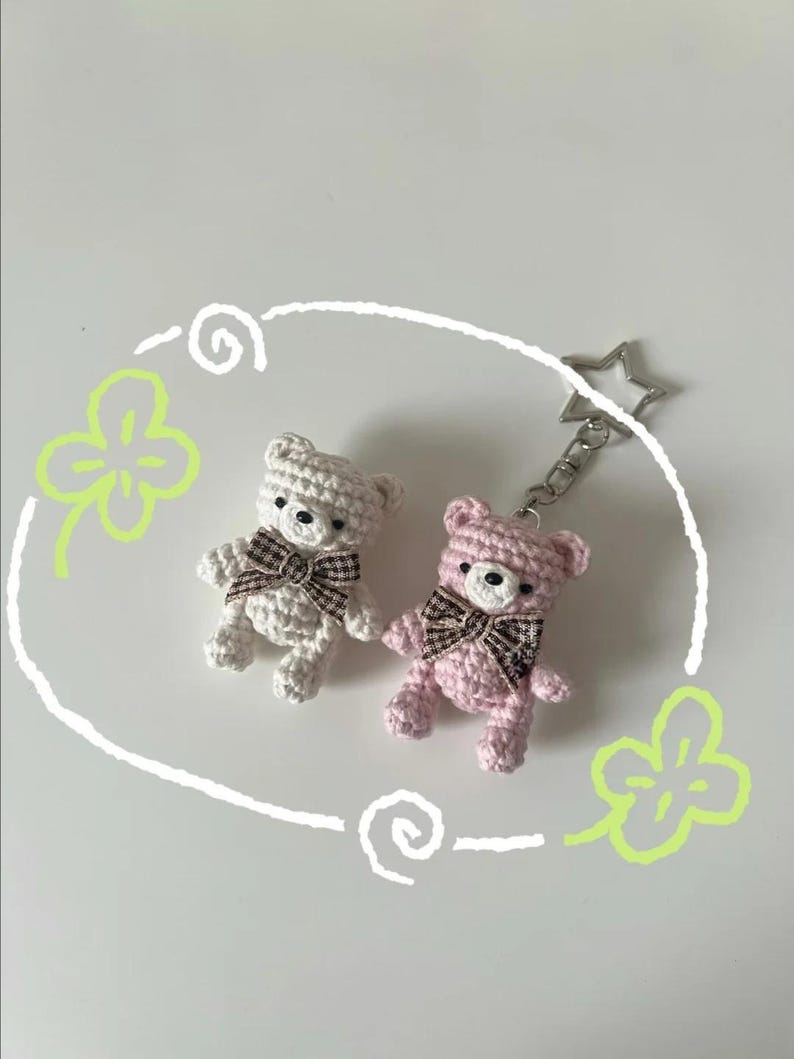 Puede incluir: Dos llaveros de osos de peluche de ganchillo. Uno es blanco, el otro rosa. Ambos tienen lazos a cuadros marrones y ojos negros. Un llavero plateado en forma de estrella est&aacute; unido al oso rosa. Los osos est&aacute;n sobre un fondo blanco con elementos decorativos verdes y blancos.