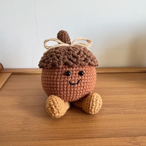 Op de afbeelding: Een gehaakte eikelpop met een lachend gezicht. De eikel heeft een bruine dop en een bruin lichaam met kleine bruine benen. Bovenop de eikel is een klein beige strikje geknoopt.