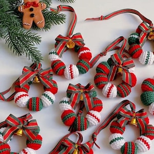 May include: Crocheted Christmas wreath decorations. Each wreath is made of red, white, and green yarn balls. They are adorned with red and green striped bows and small gold bells. A gingerbread man biscuit and pine branches are in the background.