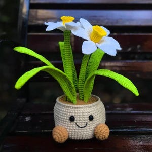 May include: A crocheted daffodil plant in a smiling pot. The white and yellow flowers have green stems and leaves. The pot is beige with a smiling face and two brown ball feet. The plant is sitting on a wooden bench.