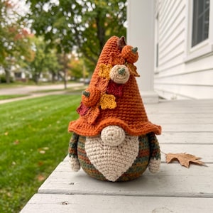 May include: A crochet gnome figurine featuring a tall, orange pointed hat decorated with small crocheted pumpkins and autumn-coloured leaves. The gnome has a round cream-coloured nose, a textured cream beard, and a body with horizontal stripes.