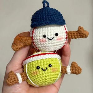 Puede incluir: Dos juguetes de ganchillo con temática deportiva. Uno es una pelota de béisbol con gorra azul, el otro una pelota de tenis con una diadema blanca y roja. Ambos tienen caras sonrientes y brazos y piernas marrones.