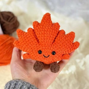 Pode incluir: Um brinquedo de pelúcia em forma de folha de bordo feito de crochê em laranja brilhante com uma superfície de fio texturizado. A folha apresenta dois pequenos olhos de contas pretas e um sorriso preto bordado, com dois pés de crochê castanhos.
