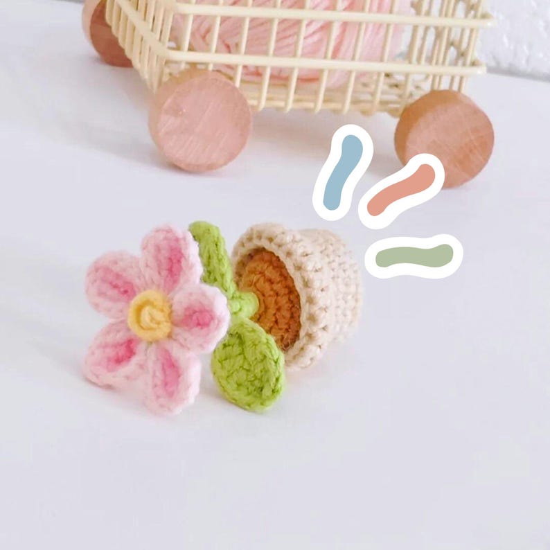 May include: Crocheted flower and potted plant. The flower is pink with a yellow centre. The potted plant is beige with an orange interior and green leaves. A small wooden cart with pink yarn is in the background.