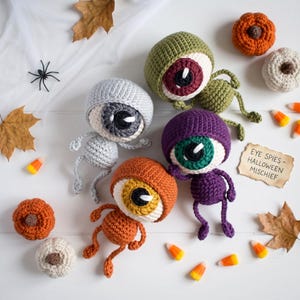 May include: Four crocheted eyeball creatures in gray, olive green, purple, and orange, each with a large iris and pupil. A small paper sign reads EYE SPIES - HALLOWEEN MISCHIEF. The scene includes small crocheted pumpkins, candy corn, and leaves.