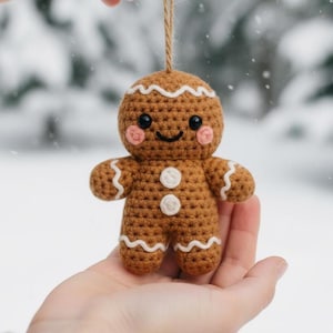 Könnte beinhalten: Handgefertigter Lebkuchenmann-Weihnachtsschmuck aus Häkelarbeit. Die braune Figur hat weiße Zuckergussdetails, rosige Wangen und ein lächelndes Gesicht. Zwei weiße Knöpfe zieren die Vorderseite. Zum Aufhängen ist eine Schnur befestigt.