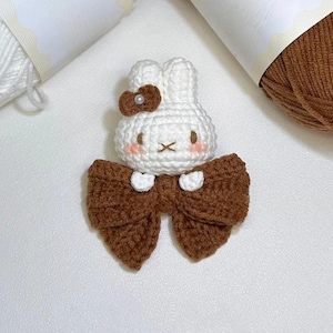 May include: A handmade crochet bunny with a brown bow and a small brown bow on its head. The bunny has a white body, pink cheeks, and a small pearl accent on the head bow. The bunny is a cute, whimsical design.