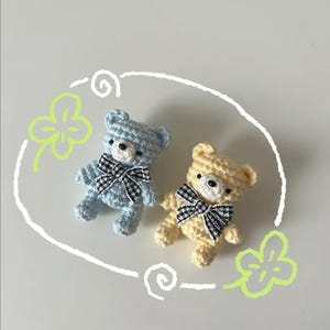 Puede incluir: Dos peque&ntilde;os osos de peluche de ganchillo. Uno es azul claro, el otro es amarillo. Ambos tienen lazos a cuadros blancos y negros. Los osos est&aacute;n rodeados por un garabato blanco y dibujos de tr&eacute;boles verdes sobre un fondo blanco.