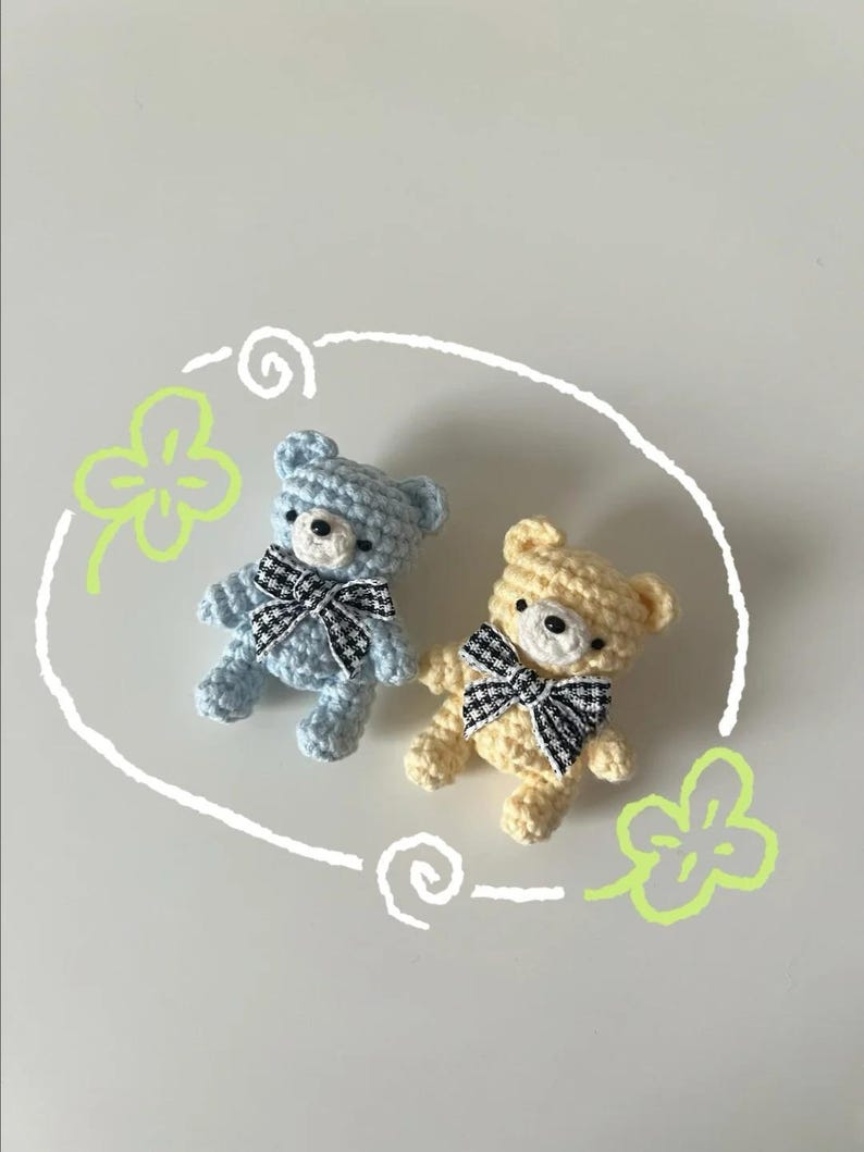 Puede incluir: Dos peque&ntilde;os osos de peluche de ganchillo. Uno es azul claro, el otro es amarillo. Ambos tienen lazos a cuadros blancos y negros. Los osos est&aacute;n rodeados por un garabato blanco y dibujos de tr&eacute;boles verdes sobre un fondo blanco.