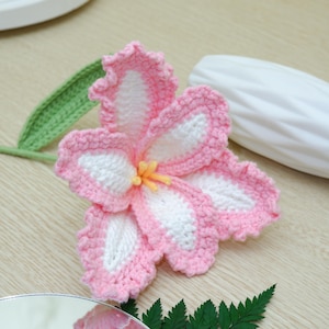 Puede incluir: Una flor de ganchillo hecha a mano en rosa y blanco, con estambres amarillos. La flor tiene un borde festoneado y está colocada sobre una superficie de madera clara. Se adjuntan una hoja y un tallo verdes de ganchillo.