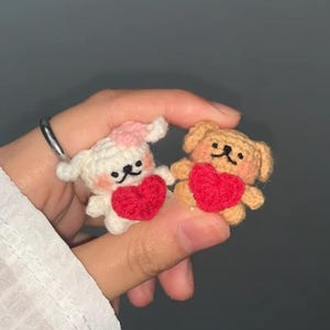 May include: Two small, crocheted animal figures: a white bunny with a pink flower and a tan bear, each holding a red heart. The figures have black embroidered facial features. The bunny and bear are handmade and likely intended as decorative items.