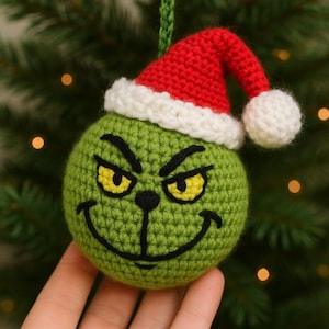 May include: A green crocheted Christmas ornament featuring the Grinch's face, complete with a red Santa hat with a white trim and a white pom-pom. The ornament has yellow eyes and a black stitched smile. The ornament is hanging from a green crocheted loop.