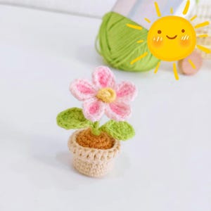 May include: A crocheted flower in a small pot. The flower has pink petals with white edges and a yellow centre. The pot is beige, and the leaves are green. The craft project is made of yarn.