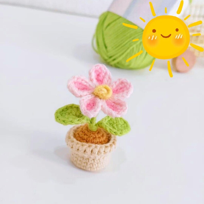 May include: A crocheted flower in a small pot. The flower has pink petals with white edges and a yellow centre. The pot is beige, and the leaves are green. The craft project is made of yarn.