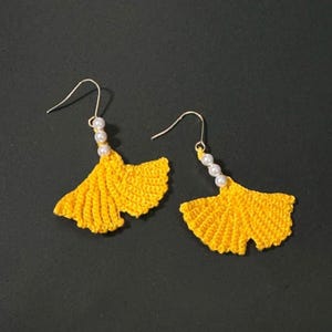 May include: A pair of yellow crochet earrings, each shaped like a ginkgo leaf. They have gold-coloured hooks and three small white beads. The earrings are displayed against a dark background.
