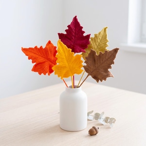 May include: A white ceramic vase containing five crocheted leaves in autumn colors including deep red, bright orange, golden yellow, mustard yellow, and brown. Each leaf is attached to a thin stem, creating a decorative arrangement of foliage.