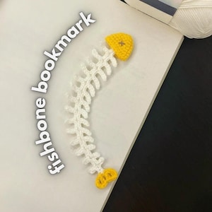 以下が含まれることがあります： 手作りの魚の骨の形をしたかぎ針編みのブックマーク。ブックマークは白で、両端に黄色のアクセントが付いています。「fishbone bookmark」というテキストが画像の左側に印刷されています。