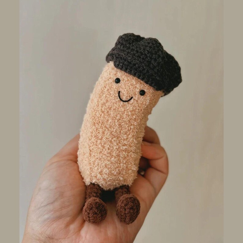 May include: A plush, beige-colored mushroom-shaped toy with a dark brown cap and small brown feet. The toy has a smiling face with black eyes and a curved mouth. The toy is held in a hand.