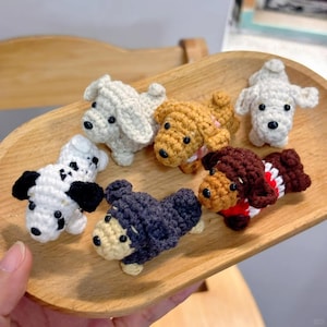 Può includere: Una collezione di figurine di cani all'uncinetto fatte a mano in vari colori, tra cui bianco, marrone e nero. I cani sono esposti su un vassoio di legno, mostrando i loro dettagli e i loro design unici. Ogni cane ha un'espressione distinta.