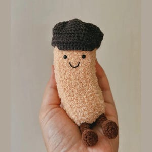 May include: A plush, smiling mushroom toy with a brown cap and small brown legs. The body is a soft, beige color. The face has a simple, stitched smile and two small black eyes. A whimsical, handmade item.