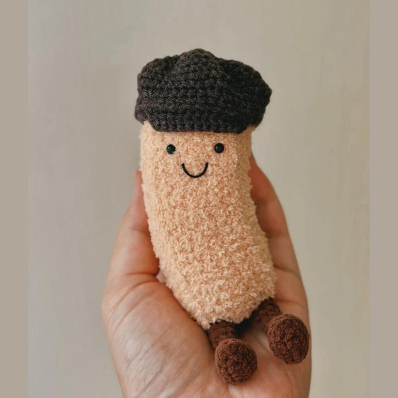 May include: A plush, smiling mushroom toy with a brown cap and small brown legs. The body is a soft, beige color. The face has a simple, stitched smile and two small black eyes. A whimsical, handmade item.
