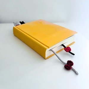 K&ouml;nnte beinhalten: Ein gelbes Hardcover-Buch mit drei handgefertigten H&auml;kel-Lesezeichen. Ein Lesezeichen ist grau mit einer bordeauxfarbenen Blume, ein anderes ist schwarz mit einer bordeauxfarbenen Blume, und das dritte ist ein Katzen-Lesezeichen. Das Buch liegt auf einer wei&szlig;en Oberfl&auml;che.