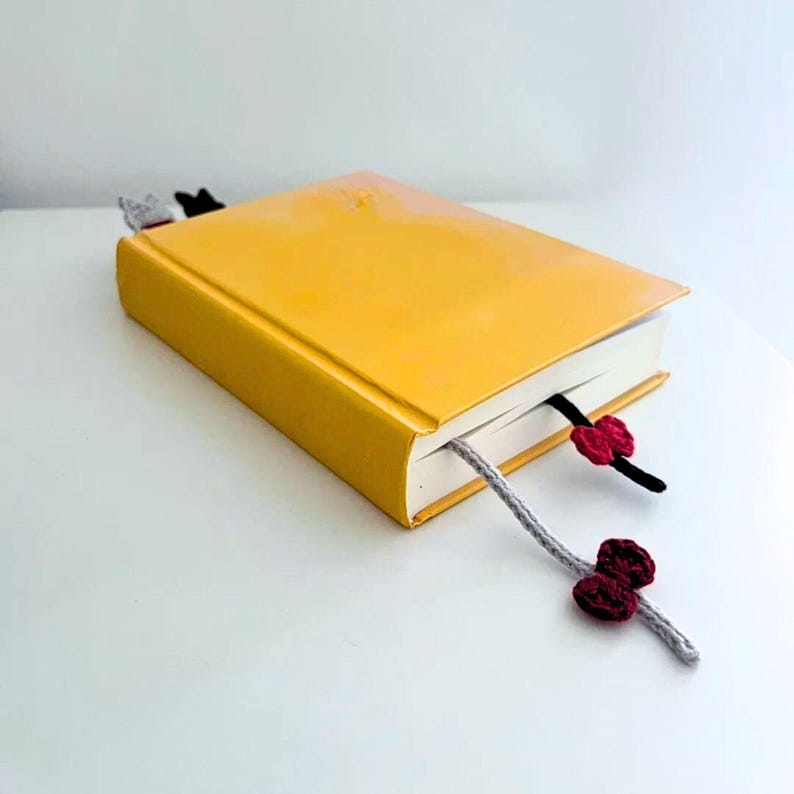 K&ouml;nnte beinhalten: Ein gelbes Hardcover-Buch mit drei handgefertigten H&auml;kel-Lesezeichen. Ein Lesezeichen ist grau mit einer bordeauxfarbenen Blume, ein anderes ist schwarz mit einer bordeauxfarbenen Blume, und das dritte ist ein Katzen-Lesezeichen. Das Buch liegt auf einer wei&szlig;en Oberfl&auml;che.