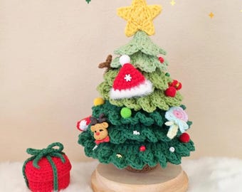 May include: A handcrafted crochet Christmas tree with a yellow star topper, adorned with miniature ornaments and a Santa hat. A small red gift box with green ribbon sits beside the tree. The tree is displayed on a wooden base.
