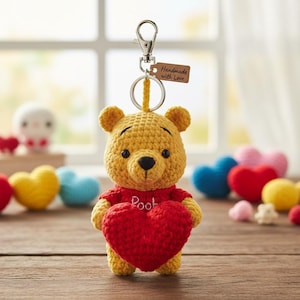 Puede incluir: Llavero de Winnie the Pooh hecho a mano, tejido a ganchillo con hilo amarillo y rojo. El oso sostiene un corazón rojo y lleva una camisa roja con "Pooh" bordado. Una pequeña etiqueta dice "Handmade with Love".