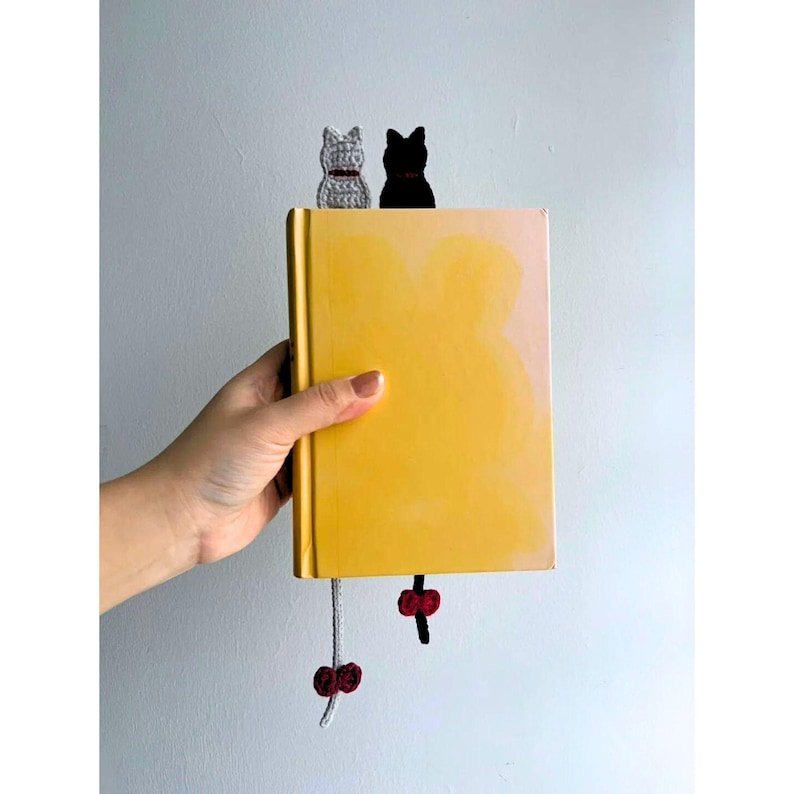 K&ouml;nnte beinhalten: Ein gelbes Buch mit zwei katzenf&ouml;rmigen Lesezeichen. Eines ist grau geh&auml;kelt mit rotem Kragen, das andere ist schwarz. Die Lesezeichen h&auml;ngen mit roten Akzenten von der Ober- und Unterseite des Buches.