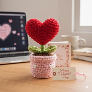 May include: A crocheted heart-shaped flower in a pink pot with white heart accents. The flower has a red heart bloom, green leaves, and a brown base. A tag that says "Happy Valentine's Day" is attached with a pink ribbon. A laptop and a card are in the background.