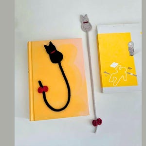K&ouml;nnte beinhalten: Zwei handgefertigte Katzen-Lesezeichen aus H&auml;kelarbeit. Eines ist schwarz mit rotem Kragen und Schleife, das andere grau mit rotem Kragen und Schleife. Die Lesezeichen liegen auf gelben B&uuml;chern.