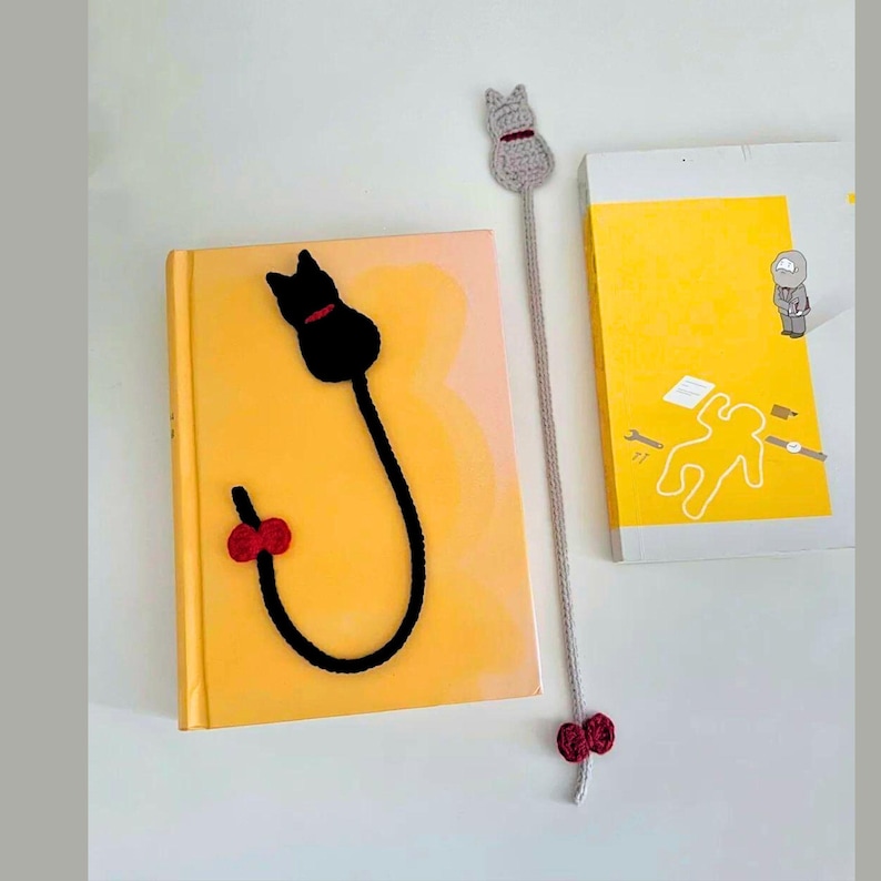 K&ouml;nnte beinhalten: Zwei handgefertigte Katzen-Lesezeichen aus H&auml;kelarbeit. Eines ist schwarz mit rotem Kragen und Schleife, das andere grau mit rotem Kragen und Schleife. Die Lesezeichen liegen auf gelben B&uuml;chern.
