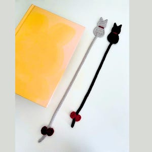 K&ouml;nnte beinhalten: Zwei handgefertigte H&auml;kel-Katzen-Lesezeichen. Eines ist grau mit rotem Kragen und Schleife, das andere ist schwarz mit rotem Kragen und Schleife. Die Lesezeichen liegen neben einem gelben Buch.