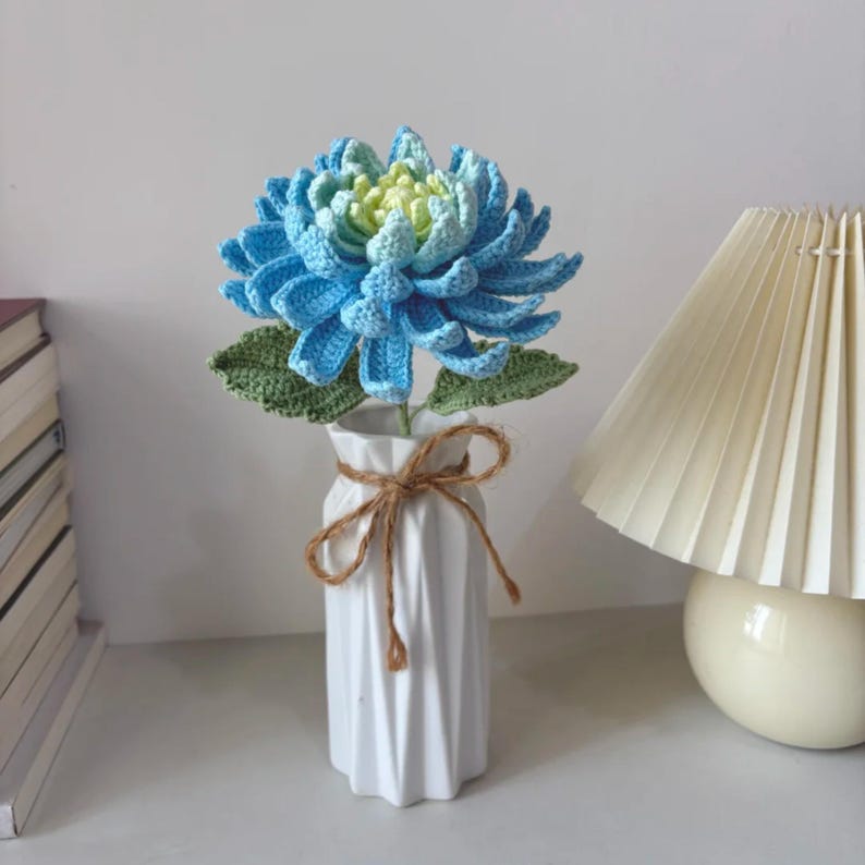 May include: A handmade crochet flower in a white vase. The flower is a vibrant blue with a gradient of yellow-green in the centre. Green leaves are visible. The vase is tied with a brown twine bow. A lamp is in the background.