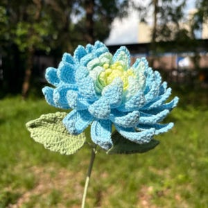 Könnte beinhalten: Eine handgefertigte Häkelblume in Blau- und Gelbtönen mit grünen Blättern und Stiel. Die Blume hat ein detailliertes, strukturiertes Aussehen, das die komplizierte Häkelarbeit zeigt. Der Hintergrund ist unscharf.