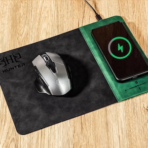 Può includere: Un tappetino per mouse con ricarica wireless nero e verde con un mouse argentato e uno smartphone. Il telefono mostra un simbolo di ricarica. Il tappetino ha la scritta "HUNTER" e le iniziali "SH2".