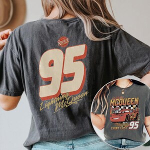 Op de afbeelding: Donkergrijze t-shirt met een grote "95" afbeelding in rood en cr&egrave;me, met de tekst "Lightning McQueen". De shirt heeft ook een kleinere afbeelding van Lightning McQueen met de tekst "Think Fast".