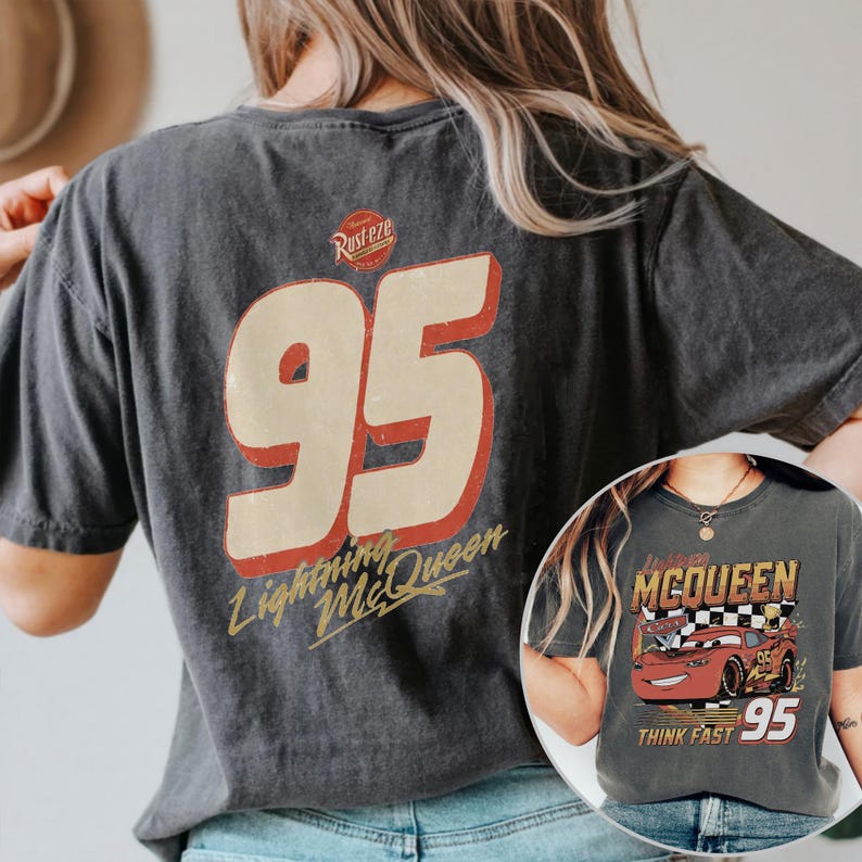 Op de afbeelding: Donkergrijze t-shirt met een grote "95" afbeelding in rood en cr&egrave;me, met de tekst "Lightning McQueen". De shirt heeft ook een kleinere afbeelding van Lightning McQueen met de tekst "Think Fast".