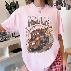 Pu&ograve; includere: T-shirt rosa chiaro con una grafica di Cricchetto del film Cars. La grafica include la scritta "MATER" sopra il carro attrezzi dei cartoni animati. Il design &egrave; in stile cartone animato con fulmini e attrezzi.