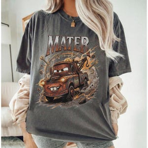 Pu&ograve; includere: T-shirt grigio scuro con un'immagine a cartoni animati di Mater del film Cars. La grafica include la parola "MATER" in arancione e marrone, con fulmini e strumenti che circondano il personaggio. La maglietta &egrave; uno stile unisex.